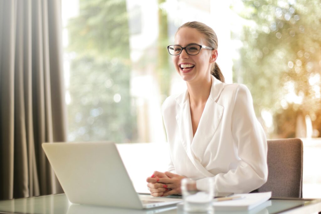 Cheerful businesswoman in glasses working on a laptop, in a bright and modern office setting.